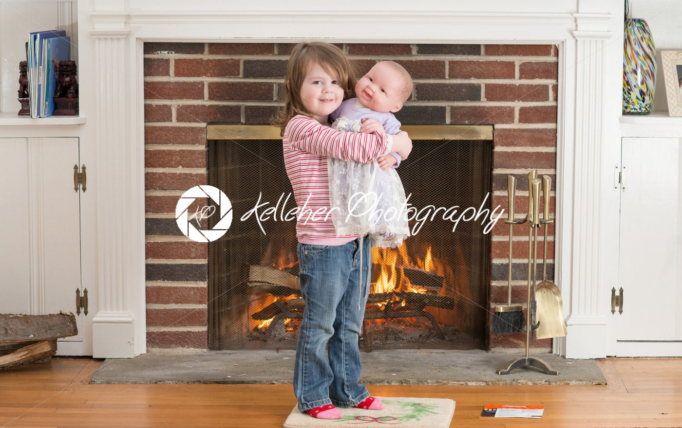 Portrait of a young smiling girl hold a baby doll in front of a fireplace dressed for valentine’s day - Kelleher Photography Store