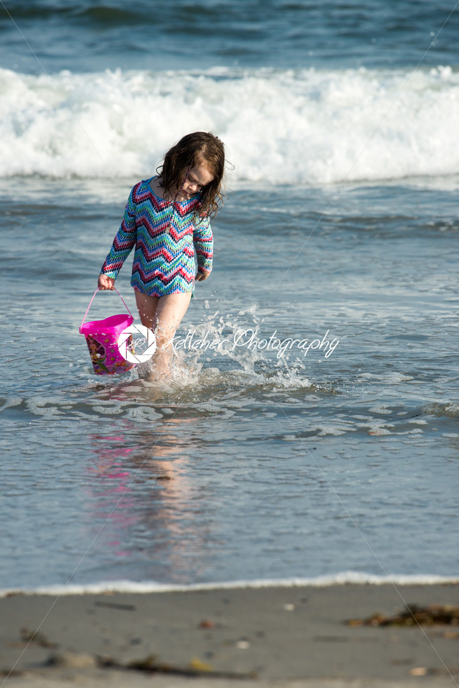 Young cute little girl playing at the seaside carrying a red bucket at the edge of the surf on a sandy beach in summer sunshine - Kelleher Photography Store