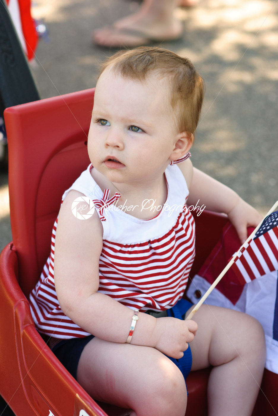 Young girl holding an American flag and riding in red wagon having fun in the park for July Fourth - Kelleher Photography Store