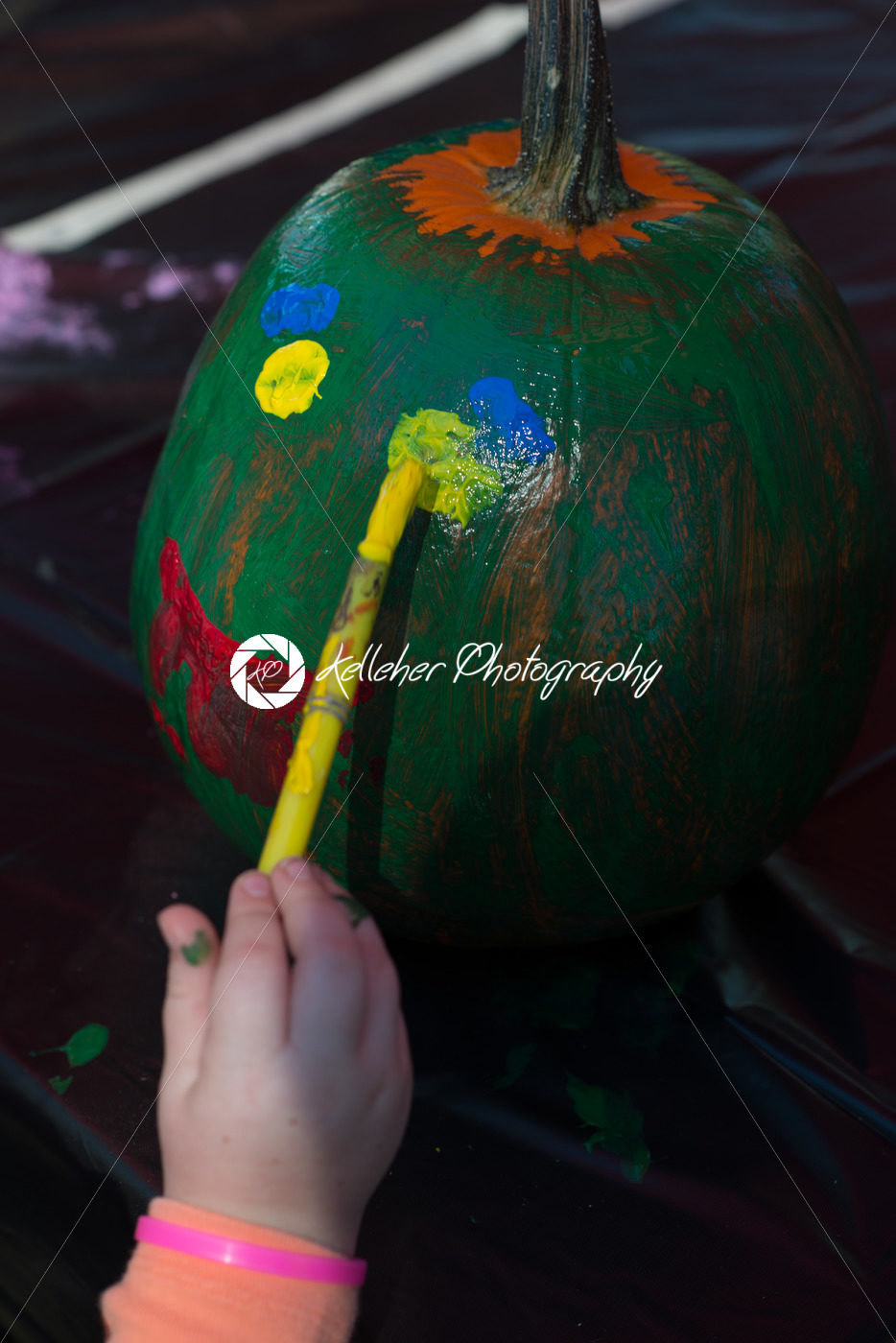 Happy little kid at harvest festival, painting a face of colors green, yellow and red on a pumpkin. - Kelleher Photography Store