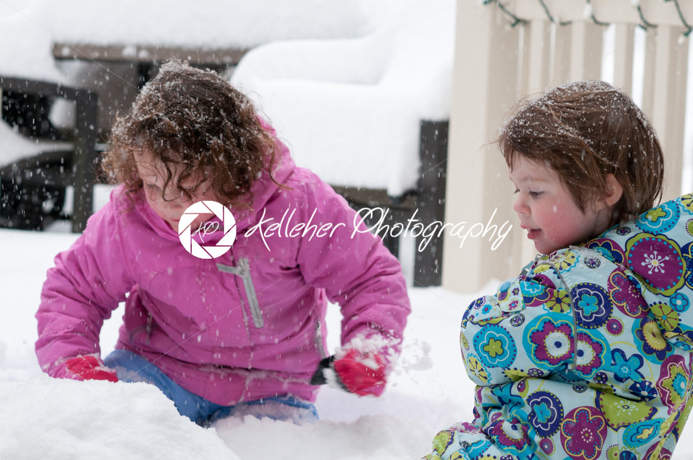 Happy toddler sibling girls in warm coat and knitted hat tossing up snow and having a fun in the winter outside, outdoor portrait - Kelleher Photography Store